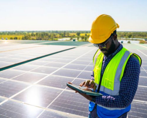 Professional African man engineer using digital tablet maintaining solar cell panels on building rooftop. Male technician working outdoor on ecological solar farm construction. Renewable clean energy technology concept